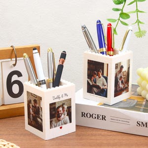 Puede incluir: Dos portalápices blancos con fotos, cada uno con bolígrafos de varios colores. Un soporte tiene el texto "Daddy & Me". Un calendario de madera muestra el número 06. Los soportes están sobre una superficie de madera.