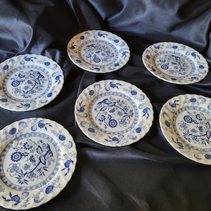 May include: Six white and blue floral patterned plates with scalloped edges. The plates feature a central floral design and a border of smaller floral and fruit motifs. The plates are arranged on a dark blue surface.