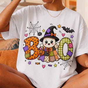 May include: A white t-shirt featuring a colourful graphic of a ghost wearing a witch hat, holding a pumpkin bucket, and surrounded by the text BOO. The design includes patchwork patterns, spiderwebs, bats, stars, hearts, and candy icons.