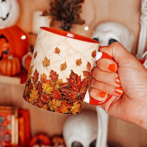 May include: A ceramic mug with a cream-colored background featuring a pattern of falling autumn leaves in shades of orange, red, and yellow. The mug has a bright orange interior and a matching orange handle, with a subtle spiderweb design.