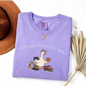 May include: A lilac Comfort Colors long-sleeve shirt with the phrase "outnumbered by boys" printed on the front. The shirt features a cartoon goose wearing a hat and holding a basketball, with a toy truck and excavator. A gold necklace is also visible.