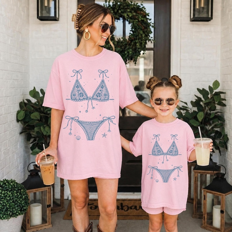 May include: Two light pink t-shirts featuring a blue graphic print of a bikini set with floral lace details, small seashells, and starfish. The shirts have a crew neckline and short sleeves, showcasing a matching design for two different sizes.