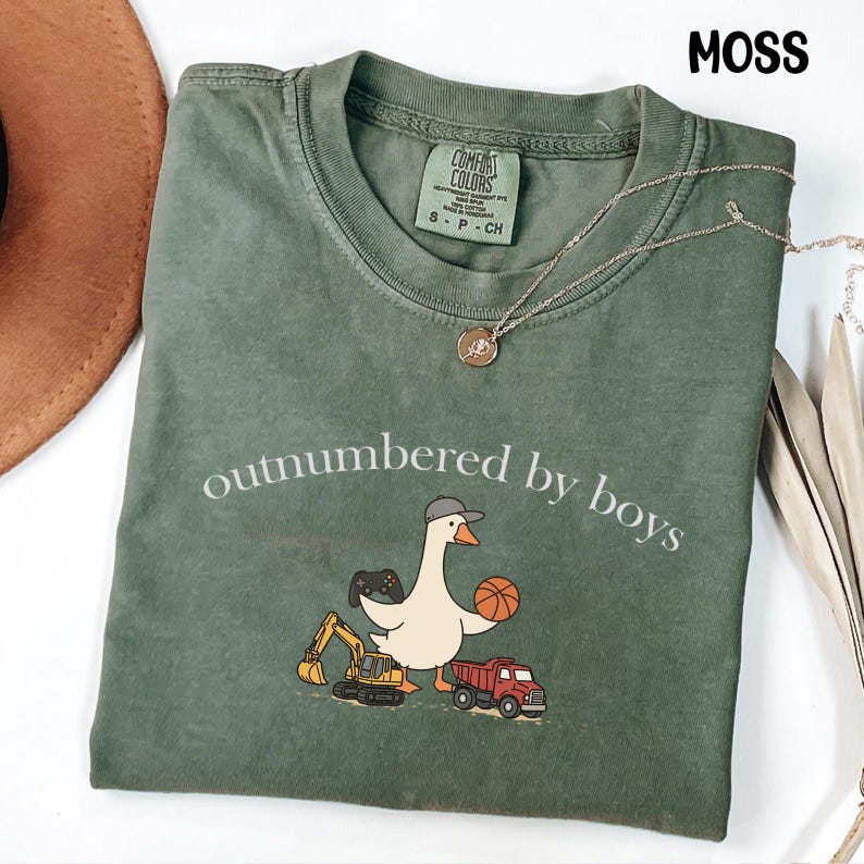 May include: A moss green t-shirt with the phrase "outnumbered by boys" printed on the front. The shirt features a cartoon goose wearing a hat and holding a basketball, with a truck, excavator, and game controller. A gold necklace is draped on the shirt.