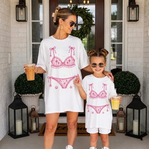 May include: Two white t-shirts featuring a pink graphic print of a bikini set with floral details, bows, and small seashell and starfish accents. The design is centered on the front of each shirt, showcasing a matching aesthetic for two different sizes.