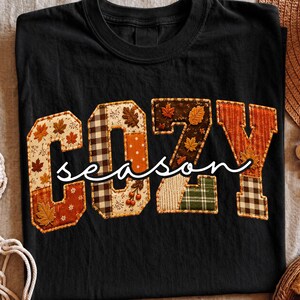 May include: A black t-shirt featuring the word COZY in large, block-style letters filled with various autumn-themed fabric patterns, including plaid, polka dots, and maple leaves. The word season is written across the center in white script.
