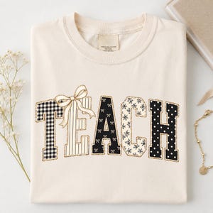 May include: A cream-coloured t-shirt featuring the word TEACH in large block letters. Each letter has a unique pattern: black and white gingham, vertical stripes, black with white bows, floral print, and black with white polka dots, plus a bow.