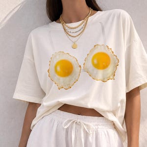 May include: An off-white short-sleeve t-shirt featuring a graphic print of two fried eggs positioned on the chest area. The shirt is paired with white drawstring shorts and a layered gold-tone chain necklace with a circular pendant.