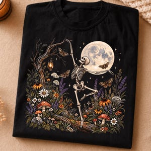 May include: A black t-shirt featuring a graphic print of a skeleton dancing in a moonlit forest. The design includes a full moon, moths, a glowing lantern hanging from a tree branch, various wildflowers, and red-capped mushrooms on the ground.