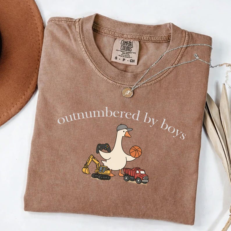 May include: Brown Comfort Colors t-shirt with the phrase "outnumbered by boys" and a cartoon goose wearing a hat, holding a basketball and a video game controller. A red truck and yellow excavator are also featured.