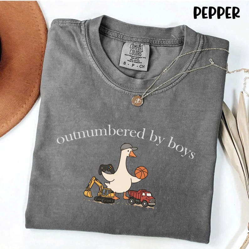 May include: A gray Comfort Colors t-shirt with the phrase "outnumbered by boys" in white. The shirt features a cartoon goose wearing a hat and holding a basketball and a video game controller. A yellow excavator and a red truck are also pictured.