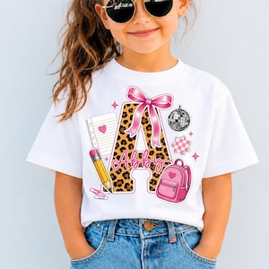Pode incluir: Uma t-shirt branca com uma letra A grande com padrão de leopardo, adornada com um laço cor-de-rosa. O design inclui um lápis amarelo, uma página de caderno com um coração, uma mochila rosa, uma bola de discoteca e o nome Abby em rosa.