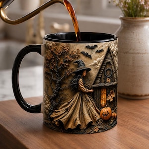 Könnte beinhalten: Ein Keramikbecher mit schwarzem Henkel und einer detaillierten Illustration einer Hexe im langen Kleid vor einem Holzhaus mit beleuchtetem Fenster, umgeben von herbstlichen Bäumen, fliegenden Fledermäusen und orangefarbenen Kürbissen.
