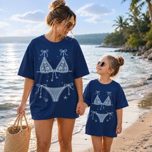 May include: Two navy blue t-shirts featuring a white graphic print of a lace bikini set. The design includes a triangle top and tie-side bottoms, accented with small star and seashell illustrations. The shirts have short sleeves and crew necks.