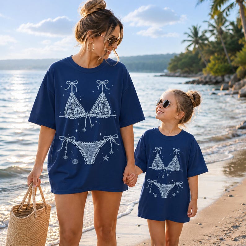 May include: Two navy blue t-shirts featuring a white graphic print of a lace bikini set. The design includes a triangle top and tie-side bottoms, accented with small star and seashell illustrations. The shirts have short sleeves and crew necks.