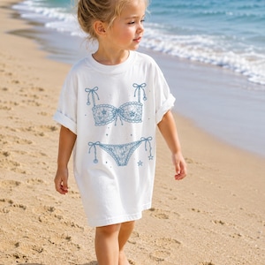 May include: A white t-shirt featuring a blue graphic print of a lace bikini set. The design includes a lace bra top with bow details and a matching lace bikini bottom with side ties, accented by small star shapes scattered around the lingerie illustration.
