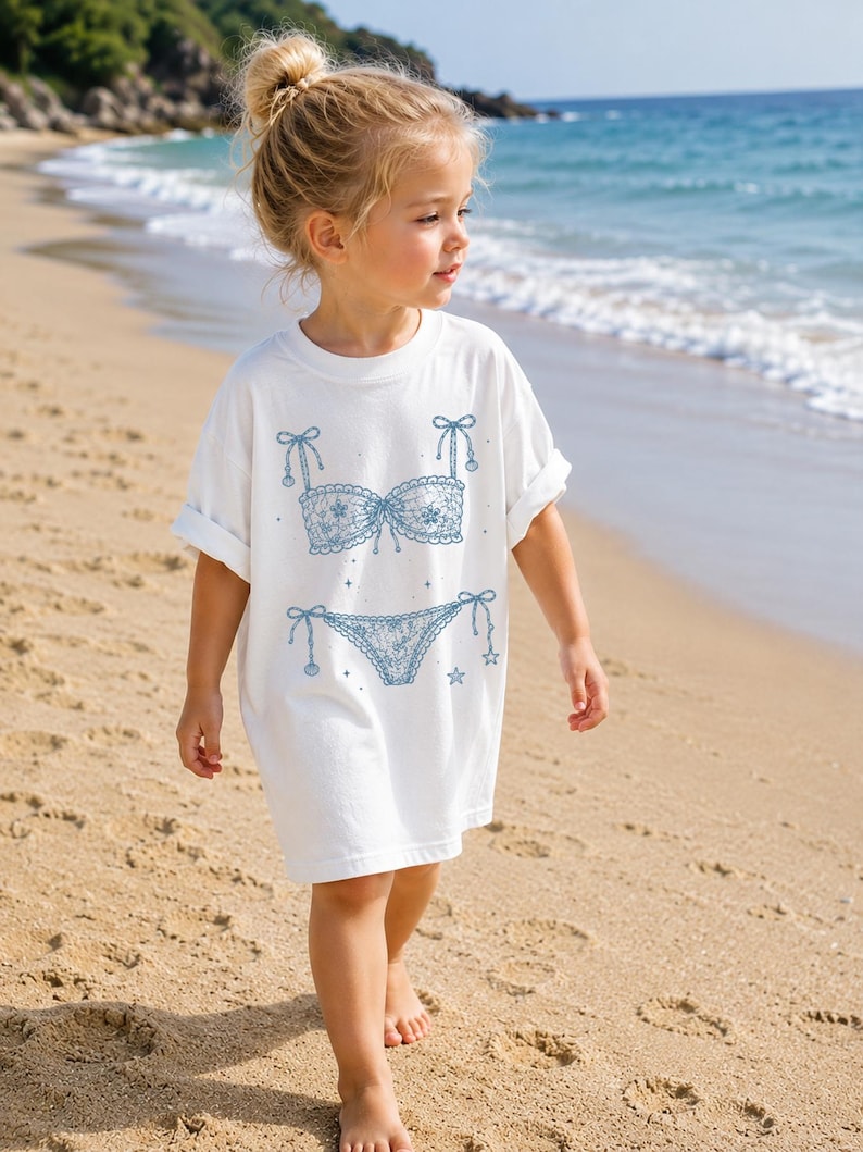 May include: A white t-shirt featuring a blue graphic print of a lace bikini set. The design includes a lace bra top with bow details and a matching lace bikini bottom with side ties, accented by small star shapes scattered around the lingerie illustration.