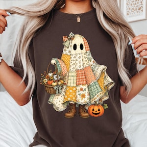 May include: A dark brown t-shirt featuring a graphic of a ghost draped in a patchwork quilt with floral and geometric patterns. The ghost wears a bow, carries a woven basket filled with autumn flowers and pumpkins, and stands beside a jack-o'-lantern.