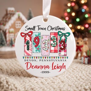 Peut inclure: Une décoration de Noël ronde en céramique blanche avec un ruban blanc. Le motif présente le texte Small Town Christmas, le nombre 1933 dans des blocs à motifs, Devon, Pennsylvania, le nom Deanna Leigh en rouge et l'année 1999.