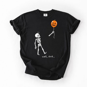 May include: A black short-sleeve t-shirt featuring a white graphic of a skeleton looking up at a detached skeletal hand holding an orange jack-o'-lantern balloon. The text "well, shit..." is printed in white below the skeleton illustration.