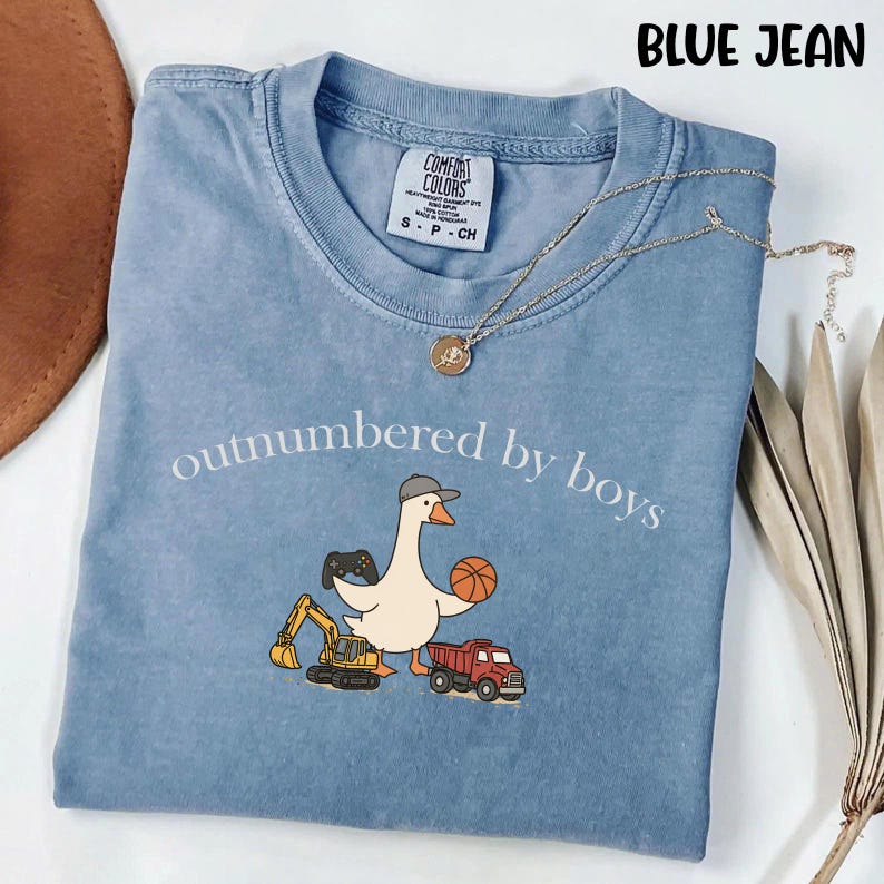 May include: Blue jean colored t-shirt with the text "outnumbered by boys" and a cartoon goose wearing a hat, holding a basketball and a video game controller. The goose is next to a toy truck and a construction vehicle. A gold necklace is on top of the shirt.