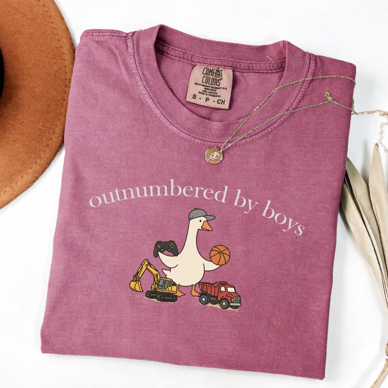 May include: A mauve Comfort Colors t-shirt with the phrase "outnumbered by boys" in white script. The shirt features a cartoon goose wearing a hat and holding a basketball and a video game controller. A small excavator and a red truck are also pictured.