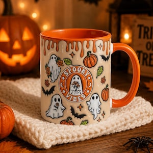 May include: A ceramic mug with an orange interior and handle, featuring a cream-colored exterior with a dripping orange glaze at the rim. The mug displays a pattern of white ghosts, orange pumpkins, and black bats, with a central circular logo reading SPOOKY VIBES.