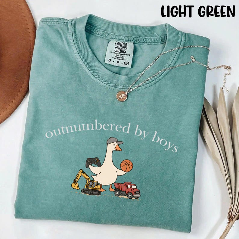 May include: Light green Comfort Colors t-shirt with the phrase "outnumbered by boys" and a cartoon goose wearing a hat, holding a basketball and a video game controller. The shirt also features a small excavator and a red truck.