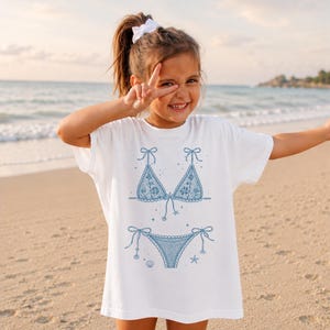 May include: A white t-shirt featuring a blue graphic print of a two-piece bikini set. The bikini design includes lace-like detailing, bows on the straps, and small illustrations of seashells and starfish surrounding the swimwear graphic.
