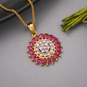 Könnte beinhalten: Goldfarbene Halskette mit einem Anhänger. Der Anhänger hat einen kreisförmigen Aufbau mit einem zentralen Cluster aus kleinen, hellvioletten Steinen, umgeben von einem Kranz aus größeren, tiefrosa, marquiseförmigen Steinen.