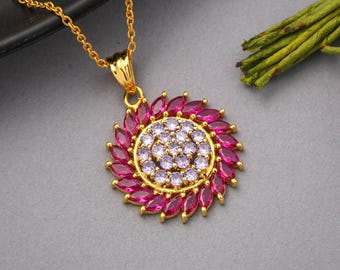 Può includere: Collana dorata con pendente circolare. Il pendente presenta un cerchio centrale di piccole pietre viola chiaro, circondato da un anello di pietre rosa scuro a forma di marquise. Anche la catena è dorata.