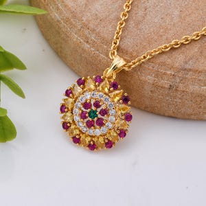 May include: A gold pendant necklace with a circular design. The pendant features a central green stone surrounded by white, yellow, and red gemstones. The necklace chain is also gold.