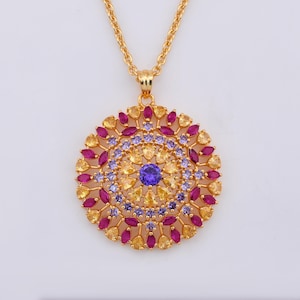 Puede incluir: Collar con colgante dorado con un diseño circular. El colgante presenta una gema azul central rodeada de círculos concéntricos de gemas amarillas, moradas y magenta. La cadena también es dorada.