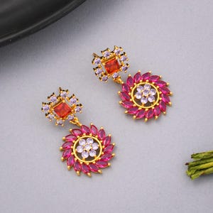 May include: A pair of gold earrings with a square, red gemstone surrounded by small, purple stones. The earrings have a floral design with pink petal-shaped stones and a centre of small, white stones.