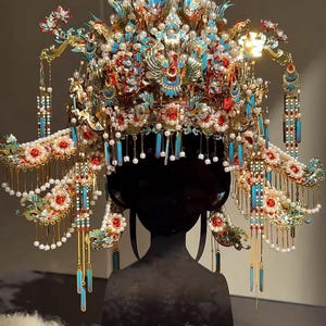 Puede incluir: Tocado ornamentado de múltiples capas y detalles intrincados. La pieza para la cabeza presenta una combinación de acentos rojos, azules y dorados, adornada con perlas y adornos colgantes. El diseño sugiere un propósito ceremonial o teatral.