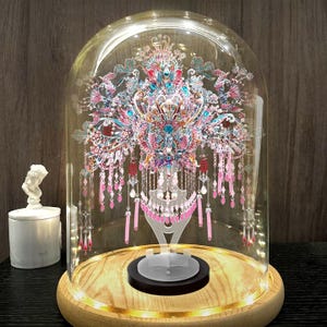Puede incluir: Una cúpula de cristal decorativa con una base de madera, que alberga una colorida e intrincada obra de arte en papel. La obra presenta tonos rosas, azules y rojos, con borlas colgantes. La base está iluminada con luces LED blancas cálidas. A la izquierda se ve una pequeña estatua blanca.