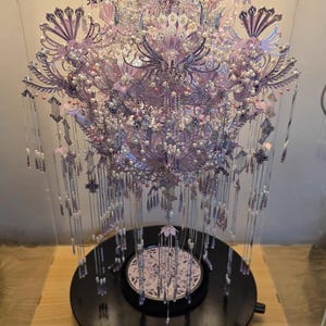 Puede incluir: Un tocado decorativo y ornamentado en una vitrina. El tocado presenta intrincados diseños en tonos lavanda, plata y blanco, con hebras de cuentas colgantes. Se encuentra sobre una base circular negra, presentado en una vitrina transparente.