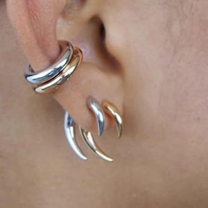 May include: Close-up of an ear adorned with two sets of earrings. One set is a double-banded ear cuff in silver and gold. The other set features three claw-shaped earrings, silver with gold tips. The jewelry is worn on a person's ear.