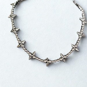Op de afbeelding: Een zilverkleurige ketting met kleine, ronde kralen en grotere, puntige spikes. De ketting is cirkelvormig, met de spikes gelijkmatig verdeeld. Het metaal heeft een gepolijste afwerking.