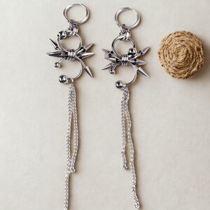May include: A pair of silver earrings with a punk rock design. Each earring has a hoop, a spiky element, and dangling chain details. A small, coiled, tan-coloured object is in the background.