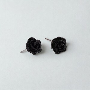 May include: A pair of black rose stud earrings. The earrings are made of a glossy material and feature detailed petals. The posts are silver-coloured. The earrings are set against a white background.