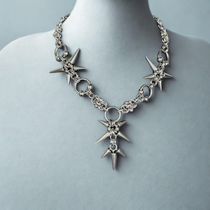 May include: Silver-toned necklace featuring a chain design with multiple spiked starburst pendants. The necklace is displayed on a mannequin, showcasing its punk-inspired aesthetic. The necklace has a bold, edgy style.