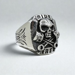 Könnte beinhalten: Silberner Ring mit Totenkopf und gekreuzten Kolben. Der Ring trägt die Inschrift "RIDE FREE". Der Ring hat eine detaillierte, strukturierte Oberfläche und ein dekoratives Design an der Seite.