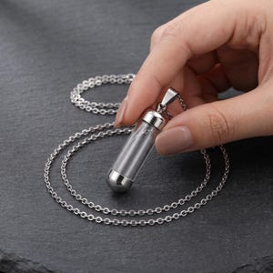 Könnte beinhalten: Eine silberfarbene Anhängerkette mit einem klaren, zylindrischen Glasfläschchen, das an beiden Enden mit polierten Metallkappen versehen ist. Der Anhänger hängt an einer feinen, silberfarbenen Kette mit kleinen, gleichmäßigen, runden Gliedern.