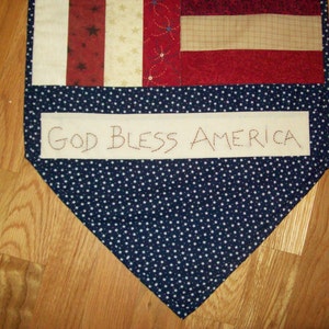 May include: A handmade fabric banner with a design inspired by the American flag. It features red and white stripes, stars, and a navy blue section with white stars. The banner displays the text "GOD BLESS AMERICA".