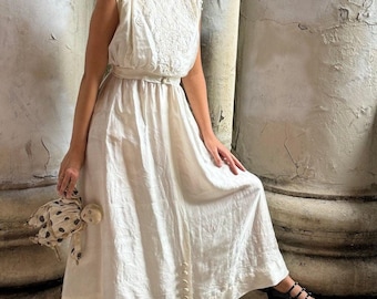 May include: A woman wearing a white sleeveless dress with lace detailing and a belt. She is standing between two pillars and holding a small spotted doll. She is wearing black shoes.