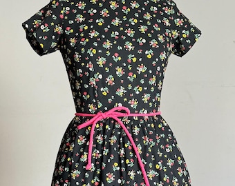 May include: A vintage black floral dress with a pink rope belt. The dress features a round neckline, short sleeves, and a ruffled hem. The fabric is covered in a pattern of small, colorful flowers.