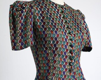 May include: A cropped, fitted, button-up blouse with short, puffed sleeves. The fabric is a black background with a repeating pattern of colorful teardrop shapes.