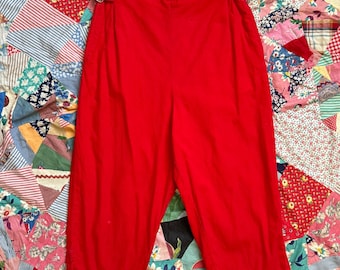 May include: Bright red trousers featuring a high-waisted design with adjustable side tabs and silver-toned metal buckles at the waist. The pants have a straight-leg cut that ends at a cropped length, showcasing a clean, solid-colored fabric finish.