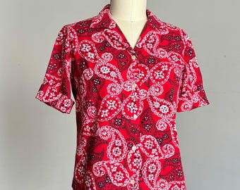 May include: A short-sleeved button-down shirt featuring a vibrant red background with a repeating white and black paisley pattern. The garment has a classic collar, a straight hemline, and light-colored buttons running down the center front.