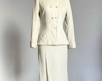 May include: A two-piece suit in a light cream color with a subtle vertical texture. The jacket features a pointed collar, long sleeves with cuffs, and a double-breasted button closure. The matching skirt is a straight, knee-length design.